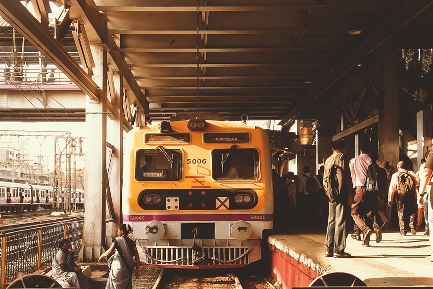 [ai] A yellow train at a busy train station platform with passengers boarding and disembarking. Other trains are visible in the background, and the station is covered with a metal roof.